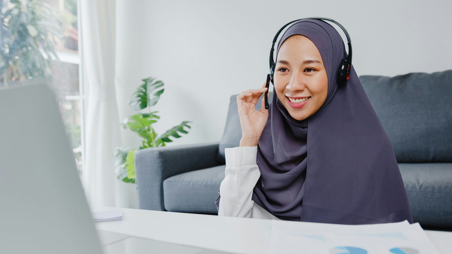 Asia muslim lady wear headphone using laptop talk to colleagues about plan in conference video call while working from home at living room. Social distancing, quarantine for corona virus prevention.